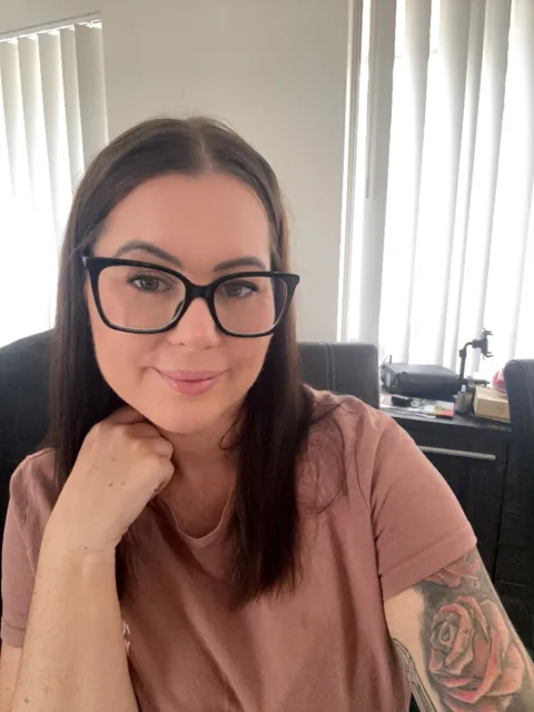 Karla Maree wearing black glasses and a dusty-pink top, resting her chin on her hand.